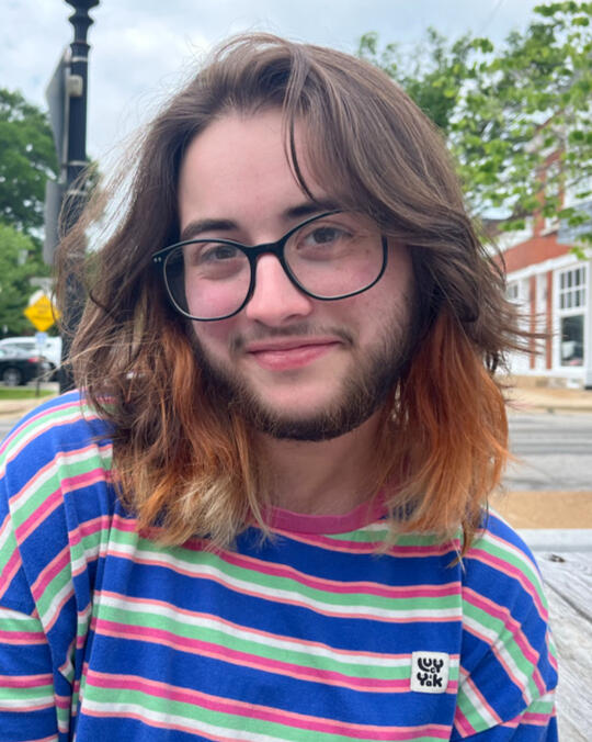 Image of Max: A white, masculine-presenting person with long dark brown hair with orange/blonde highlights, a dark brown beard, dark brown eyes, and black glasses. He is wearing a pink, blue, green, and white striped shirt from the brand Lucy & Yak.